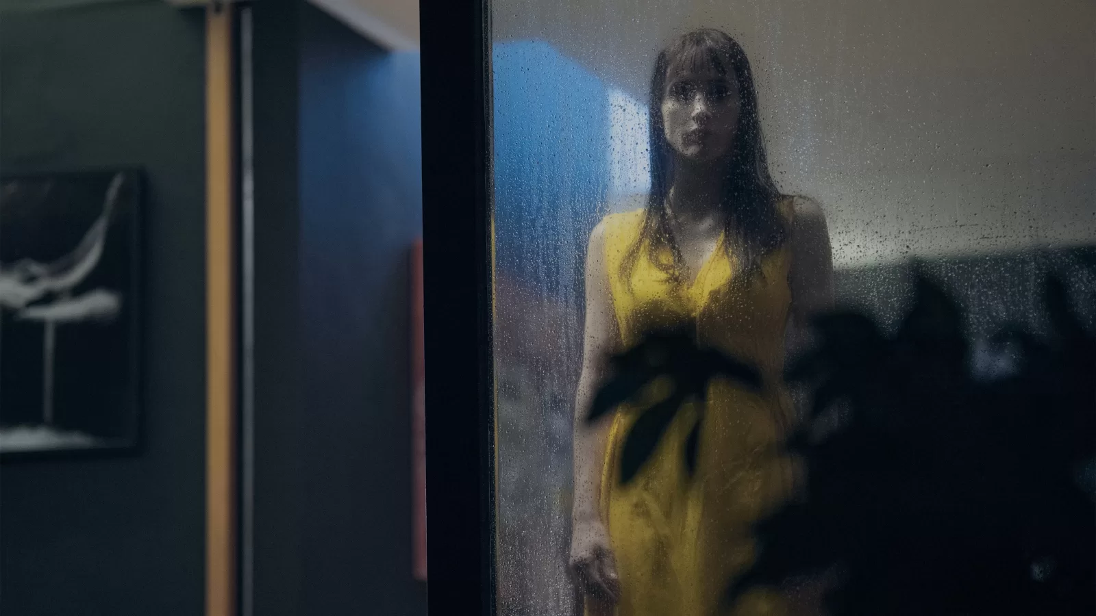 Mujer de vestido amarillo y cabello largo de pie tras un vidrio empañado en una habitación en penumbra