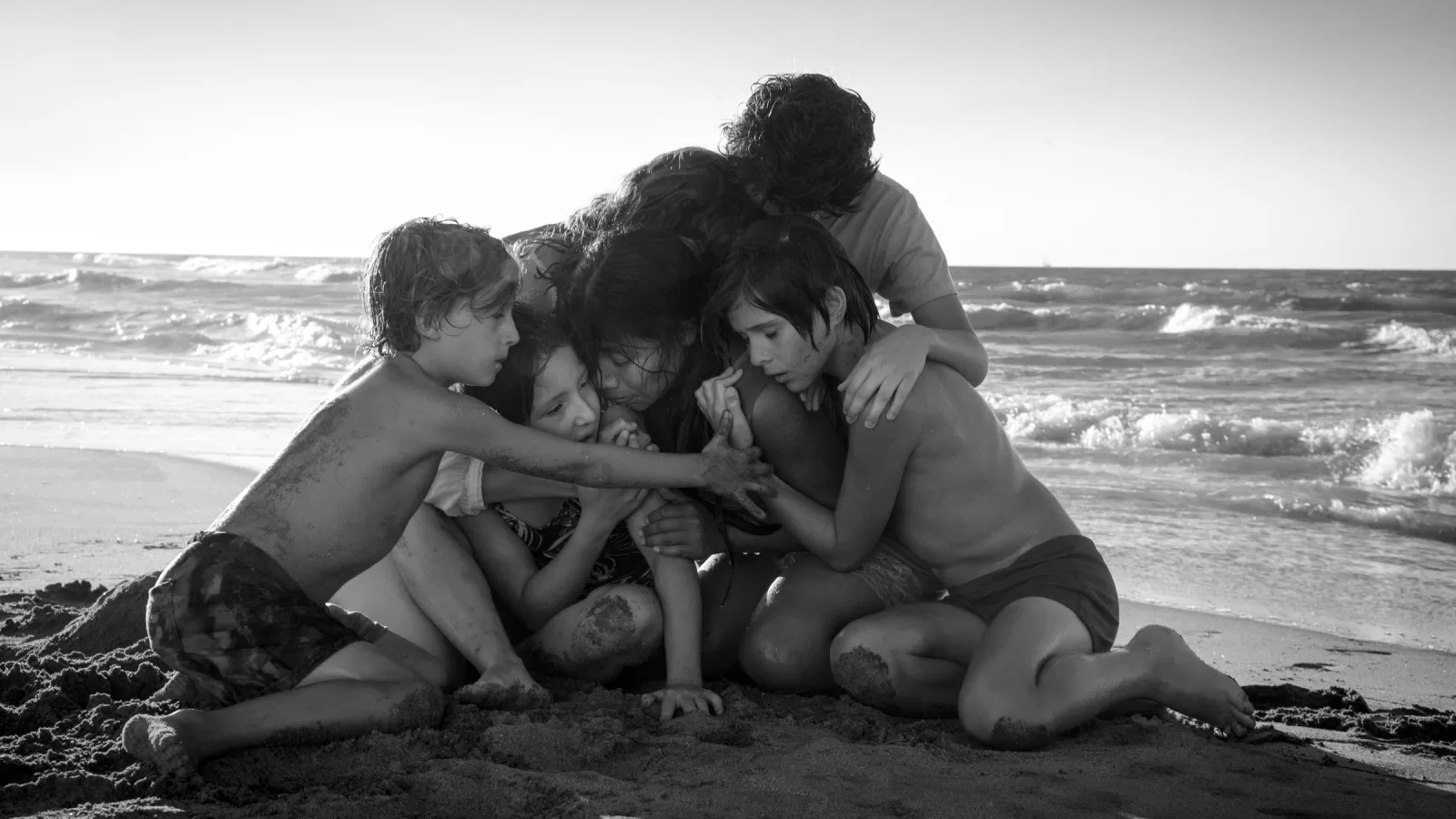 En blanco y negro, una mujer abraza a un grupo de niños empapados sentados en la arena de una playa